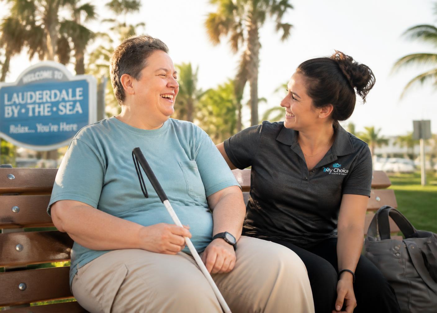 A companion caregiver chatting warmly with the adult she supports on a sunny park bench.